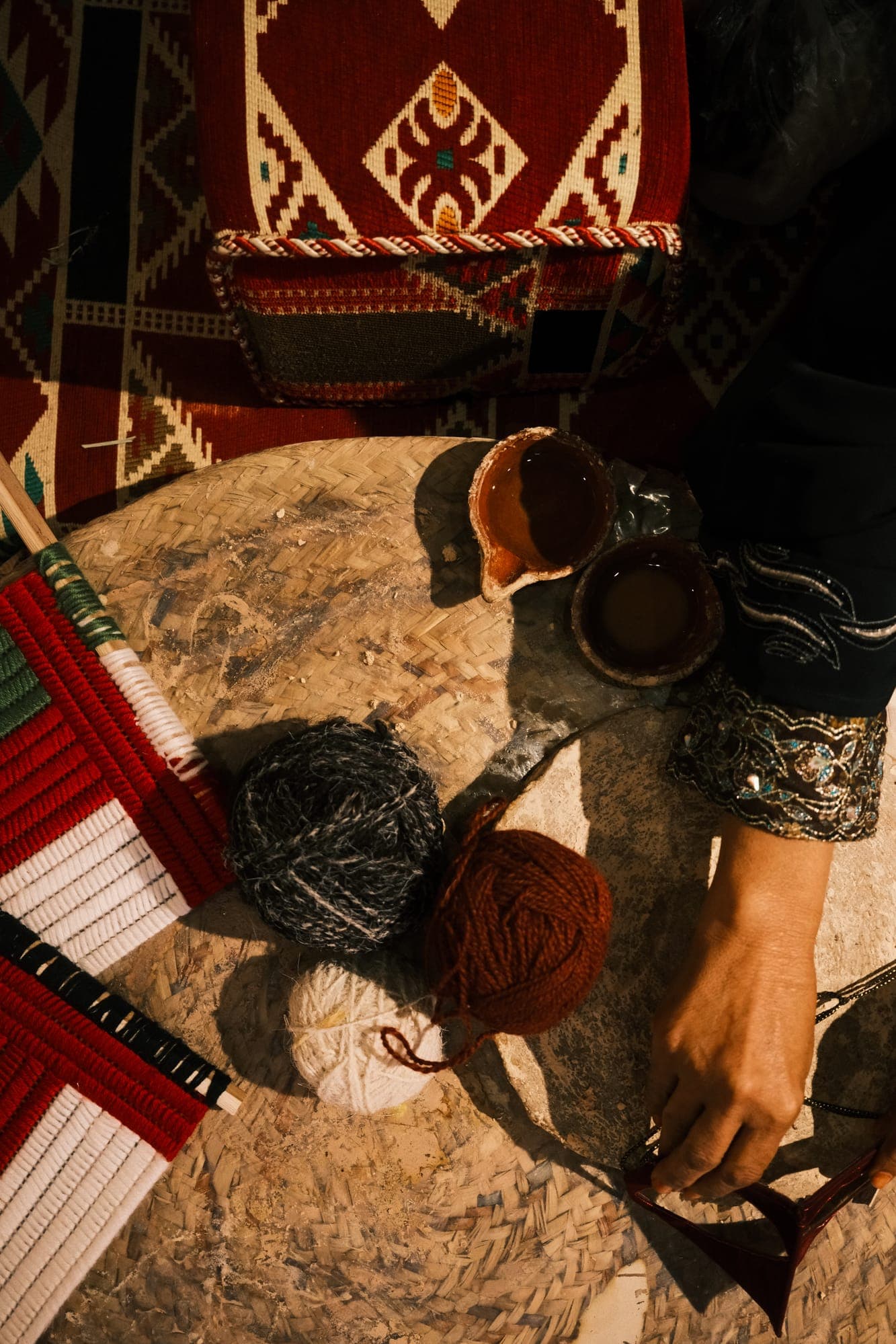 من الأعلى: كُبَب غزل، وكوبان صغيران من الفخار، ويد تمسك شريطاً داكناً فوق حصير منسوج.